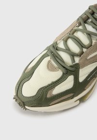 Shoe com um upper de malha bege, acentos verde oliva e detalhes em camurça texturizada. Apresenta atacadores notáveis e uma sola de borracha para aderência.