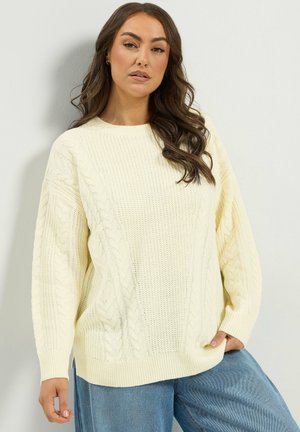 Femme aux longs cheveux bruns portant un pull crème torsadé et un jean bleu, debout devant un fond clair uni.