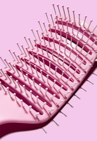 Brosse à cheveux avec une base en plastique rose et des poils blancs. Elle présente une forme courbée et une série de rangées arquées pour démêler les cheveux.