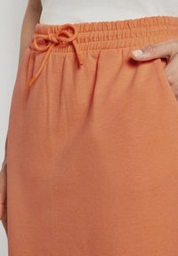 Pantalones de chándal naranja con cintura elástica y cordón. Textura suave, con bolsillos laterales y tejido fruncido en la cintura.
