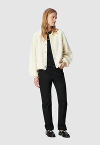 Veste en fausse fourrure blanche avec des boutons argentés, haut noir, pantalon texturé noir et chaussures noires, portée par une personne se tenant devant un fond neutre.