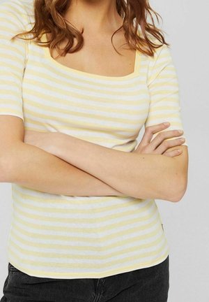 Person med korslagte arme iført kortærmet gul og hvidstribet top og sorte bukser, afskåret ved hals og talje.