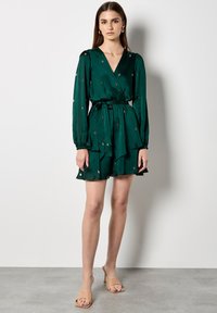 Groene wikkeljurk met lange mouwen, gerimpelde zoom en gouden bloemenborduursel. Zachte stof met een taillering en bijpassende ceintuur.