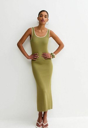 Kobieta w oliwkowozielonej, bezrękawnikowej, żeberkowanej sukience maxi z białą lamówką, kolczykach kołach, bransoletkach i białych sandałach, stojąca na tle jednolitej ściany.