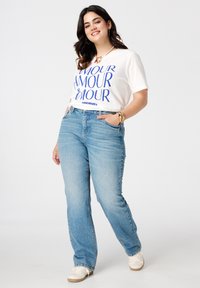 Witte t-shirt met blauwe tekst "AMOUR" en "TOUJOURS," gecombineerd met lichtblauwe spijkerbroek en witte sneakers. Model heeft een relaxte houding.