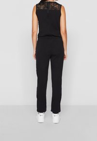 Svart ärmlös jumpsuit med spetsdetalj på ryggen, elastisk midja och rak bendesign, kombinerad med vita sneakers.