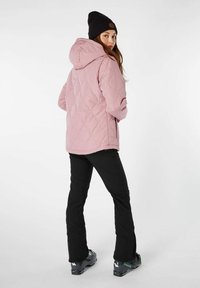 Lichtroze gewatteerde jas met capuchon, zwarte beanie en zwarte broek; heeft een gestructureerd oppervlak en een aansluitend design; gedragen met skibotten.