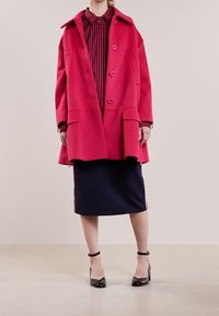 Cappotto di lana fucsia con grandi bottoni e tasche laterali, sovrapposto a una gonna navy con strisce laterali, abbinato a tacchi neri con cinturini alla caviglia.