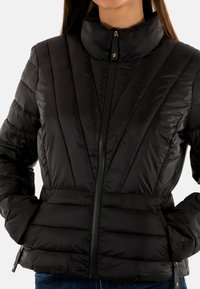 Giacca nera imbottita con colletto alto, motivo trapuntato verticale, chiusura con zip e tasche laterali con finiture in tono oro.