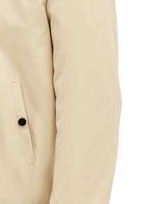 Beige pufferjack met een gladde textuur, voorzien van een zijzak met een zwarte knop. Het ontwerp is eenvoudig met een ontspannen pasvorm.