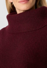 Woman wearing gold hoop earrings and a ribbed, maroon knit turtleneck sweater with a folded collar.