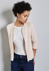 Svetle béžová prošívaná bunda s kulatým výstřihem, texturovanou tkaninou a zapínáním na zip, kombinovaná s bílou košili a tmavě modrými džíny.