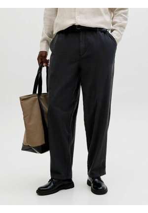 Person trägt dunkle, lockere Hosen, schwarze Schuhe, cremefarbenes Hemd, hält eine beige-schwarze Tragetasche und steht vor einem einfachen hellen Hintergrund.