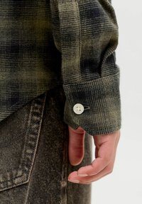 Button-up skjorte med et grønt og sort plaidmønster. Stofteksturen virker blød. Har en hvid knap ved manchetten, med denim synligt nedenfor.