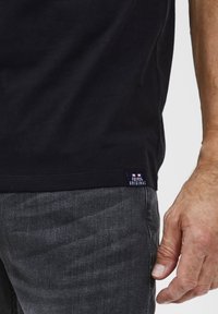 Melna kokvilnas T-kreklu ar īsām piedurknēm, apaļu kakla izgriezumu un nemanāmu emblēmu uz apakšmalas, kombinēts ar tumši pelēkiem džinsu biksēm.