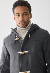 Vīrietis valkā pelēku mēteli ar pogām uz pleciem pār krēmkrāsas adītā turtleneck džemperi uz vienkārša gaismas fona.