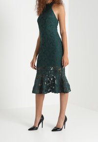 Grünes Spitzenkleid mit tailliertem Oberteil, schwingendem Saum und Halterneck; es weist florale Muster und eine halbtransparente Textur auf, kombiniert mit schwarzen Absätzen.