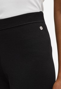 Fekete leggings texturált anyagból, bordázott derékrésszel és ezüst színű logógomb egyszerű díszítéssel az oldalon.