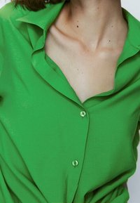 Chemise verte à boutons avec un col. Fabriquée en tissu léger, elle présente une coupe décontractée et un léger éclat. Les boutons sont de la même couleur verte.