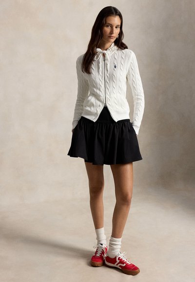 Kvinne iført hvit kabelstrikket genser med glidelås, svart kort skjørt, hvite sokker og røde joggesko, stående foran en ensfarget lys bakgrunn.