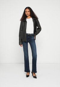 Jachetă neagră din piele cu guler ascuțit, bluză albă, blugi evazați în albastru închis și pantofi cu toc înalt negri. Textură netedă, design minimalist.