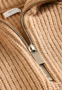 Beige gebreide stof met verticale ribbels, voorzien van een zilverkleurige metalen rits en een label met de tekst "NEXT." Textuur oppervlak, gezellige uitstraling.