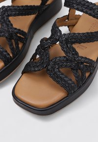 Sandalias de cuero trenzado negras con una suela plana en tono marrón. Presentan correas entrelazadas y una suela de plataforma suave.