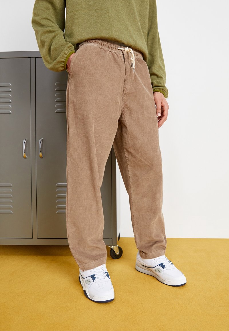 Ljusbruna kordbyxor med elastisk midja och dragstring. Bärs med vita sneakers. Grå metallskåp i bakgrunden.