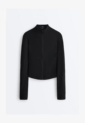 Schwarze, eng anliegende Jacke mit langen Ärmeln, Frontreißverschluss, hohem Kragen und Nahtdetails, auf hellem, einfarbigem Hintergrund präsentiert.