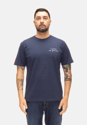 Uomo con capelli corti e scuri e barba, che indossa una maglietta blu navy con testo in italiano e jeans, in piedi su uno sfondo bianco semplice.