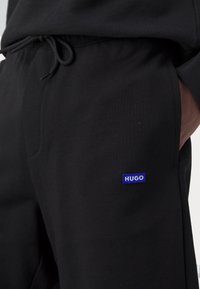 Gros plan d'un pantalon de survêtement noir avec une taille à cordon et une petite étiquette rectangulaire bleue "HUGO" sur la cuisse droite.