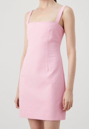 Kobieta ubrana w różową, bezrękawnikową mini sukienkę z kwadratowym dekoltem i fakturowaną tkaniną na jednolitym jasnym tle.