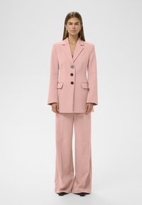 Helle rosa maßgeschneiderte Blazerjacke mit drei schwarzen Knöpfen, breiten Revers und zwei vorderen Taschen, kombiniert mit passenden, weit geschnittenen Hosen.