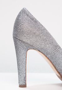 Stříbrný třpytivý textilní vysoký podpatek s špičatou špičkou a robustním blokovým podpatkem, umístěný na jednoduchém bílém pozadí.