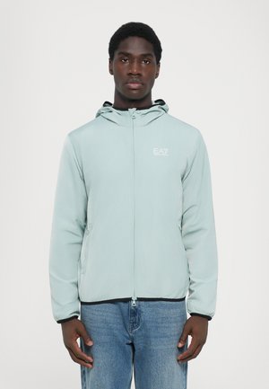 Giacca zip-up blu chiaro con cappuccio, realizzata in materiale soffice. Presenta due tasche laterali e un logo bianco sul petto.