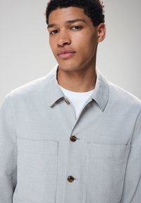 Light gray textured jacket with a collared neckline, two front pockets, and brown buttons. Worn over a white shirt.