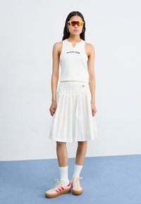 Hvit ermeløs topp med "Dickies" trykt, plisserte hvite skjørt og flerfargede platåsko med blomsterdetaljer. Klare solbriller.