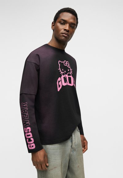 Νέος άνδρας φοράει μαύρο μακρυμάνικο μπλουζάκι με ροζ γραφικά Hello Kitty και το κείμενο "GCOSWEAR" στο μανίκι, συνδυασμένο με ανοιχτό γκρι τζιν.