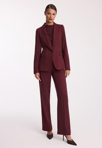 Burgundowa dopasowana garsonka z kieszonką w fasonie slim fit, z pojedynczym guzikiem i klapami w kształcie notcha, zestawiona z prostymi spodniami i szpiczastymi szpilkami.