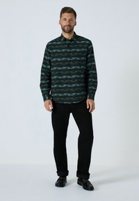 Vihreä ja musta kuvioitu nappipaitapusero, jossa on geometrinen muotoilu, pitkät hihat ja tavallinen kaulus, yhdistettynä mustiin housuihin ja kenkiin.