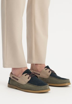 Zweifarbige Freizeitschuhe mit dunkelblauem und hellbraunem Wildleder, braunen Schnürsenkeln und einer strukturierten Gummisohle, getragen mit beigen Hosen.