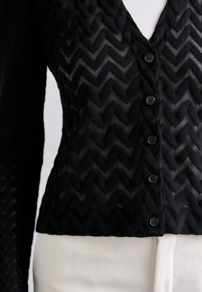 Gilet noir à boutonner avec une texture en chevrons. Présente un décolleté en V profond et six boutons sur le devant.