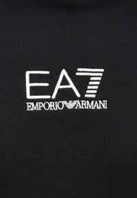 Λογότυπο EA7 Emporio Armani κεντημένο με λευκό νήμα σε μαύρο ύφασμα, με στυλιζαρισμένο '7' με οριζόντιες ρίγες.