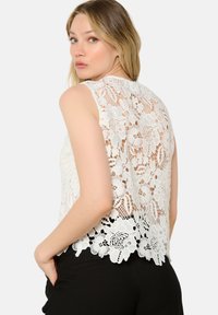 Top in pizzo bianco con motivo floreale, design senza maniche, orlo scallopato e dettaglio trasparente sulla schiena. Abbinato a pantaloni neri.