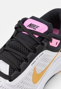 Scarpa sportiva in bianco e nero con tomaia in mesh testurizzato, swoosh Nike arancione, etichetta logo Nike rosa e lacci neri. Dettagli del Dynamic Fit System visibili.