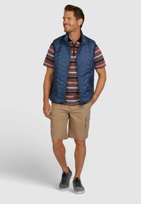 Marineblaue gesteppte Weste über einem gestreiften kurzärmligen Poloshirt. Bequeme beige Cargo-Shorts. Graue Sneakers mit schwarzen Schnürsenkeln. Schlichtes, funktionales Design.