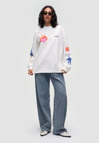Camicia bianca a maniche lunghe con stampati colorati raffiguranti un cuore infuocato, labbra e uccelli sulle maniche, abbinata a jeans blu larghi.