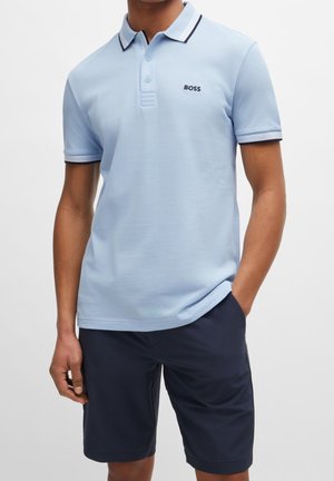 Mann trägt hellblaues Poloshirt mit kurzen Ärmeln und "BOSS"-Logo sowie dunkelblaue Shorts, eine Hand in der Tasche, vor einfarbigem Hintergrund.