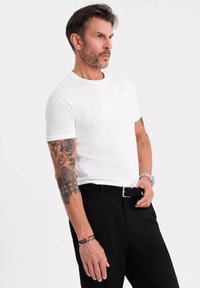 Hvid kortærmet t-shirt med rund hals, båret med sorte bukser. Har detaljerede tatoveringer på armene og et sølvur med et sort rem.