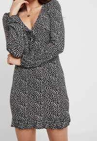 Czarna sukienka z białymi wzorami kwiatowymi, dekolt w serek, długie rękawy i falbaniasty dół. Wykonana z lekkiego materiału o dopasowanym kształcie.